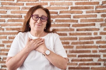 Poster - Senior woman with glasses standing over bricks wall smiling with hands on chest with closed eyes and grateful gesture on face. health concept.