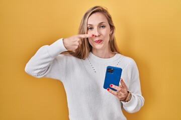 Canvas Print - Young blonde woman using smartphone typing message pointing with hand finger to face and nose, smiling cheerful. beauty concept