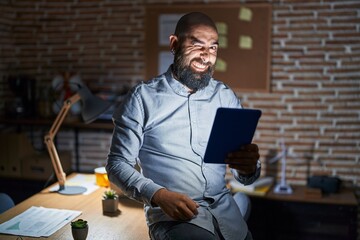 Wall Mural - Young hispanic man with beard and tattoos working at the office at night winking looking at the camera with sexy expression, cheerful and happy face.