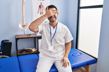 Poster - Young hispanic man with beard working at pain recovery clinic smiling and laughing with hand on face covering eyes for surprise. blind concept.