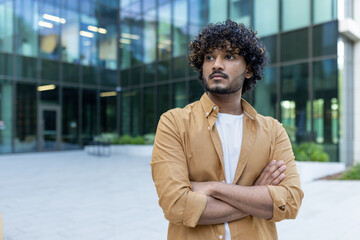 Portrait of a young Indian male programmer, developer standing with crossed arms outside the office center, seriously and confidently looking to the side