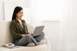 © Prostock-studio - Smiling chinese woman sitting on couch with computer