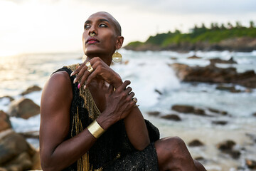 Wall Mural - Non-binary black person in luxury dress sits on rocks in ocean. Trans ethnic fashion model wearing jewelry in posh gown, poses in tropical seaside location. Divine feminine human. Diversity concept.