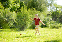 Funny Little Boy Playing With Garden Sprinkler In Sunny Backyard. Preschooler Child Laughing, Jumping And Having Fun With Spray Of Water.
