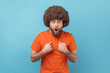 © khosrork - Portrait of man with Afro hairstyle in orange T-shirt points himself, looking with surprised expression, shocked and proud of win, unbelievable success. Indoor studio shot isolated on blue background.