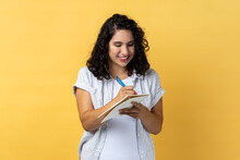 Portrait Of Smiling Satisfied Attractive Woman With Dark Wavy Hair Writing In Paper Notebook, Making Notes, Schedule Or To Do List. Indoor Studio Shot Isolated On Yellow Background.