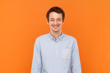 Portrait of overjoyed positive happy man standing looking at camera with toothy smile, expressing happiness, wearing light blue shirt. Indoor studio shot isolated on orange background.