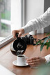 © Pixel-Shot - Woman pouring coffee from pot into cup on wooden windowsill