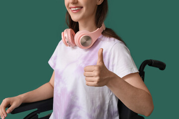 Wall Mural - Young woman in wheelchair showing thumb-up on green background, closeup