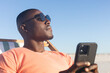 © wavebreak3 - Happy african american man in sunglasses sitting in deckchair using smartphone on sunny beach