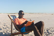 © wavebreak3 - Happy african american man in sunglasses sitting in deckchair reading book on sunny beach