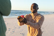 Happy african american man kneeling with ring and proposing to girlfriend on sunny beach