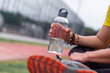 Athlete with bead bracelets holding plastic water bottle resting after training at outdoor sports arena man sitting with beverage on urban stadium closeup
