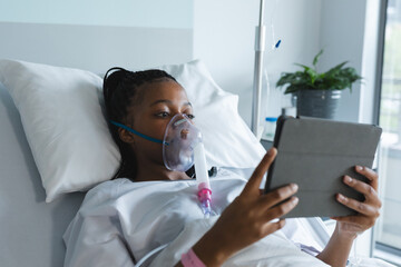 Wall Mural - African american female patient with oxygen mask, lying on bed and using tablet in hospital room