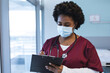 African american female doctor wearing scrubs and face mask, holding clipboard and writing