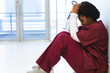 Tired african american female doctor wearing scrubs, sitting on floor in corridor at hospital