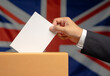 Hand voter holding ballot paper putting into the voting box at place election against the United Kingdom flag background