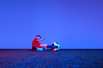 Wall Mural - Male beginner soccer player in sports uniform in action, motion with ball over dark blue background in neon. Concept of sport, health, action, ad