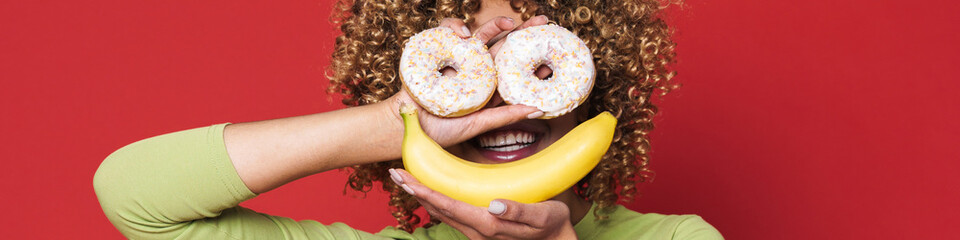 Wall Mural - Young black woman with afro curls having fun holding donuts and banana