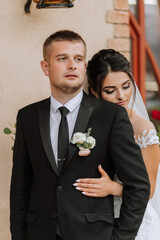 Wall Mural - A stylish couple of European newlyweds. A smiling bride in a white dress hugs the groom from behind. Groom dressed in a classic black suit, white shirt, tie. Wedding in nature