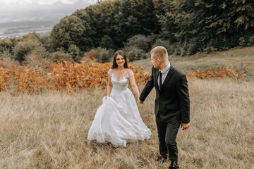 Wall Mural - A romantic fairytale couple of newlyweds are walking in a field at sunset, behind fern leaves. The bride is in a white wedding dress, the groom is in a black classic suit and a white shirt