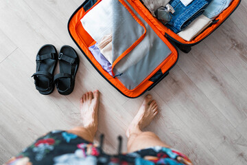 A young man is packing clothes in a suitcase in his apartment for the summer holidays. The luggage is on the floor. He checks if everything is packed. The sandals are next to the suitcase.