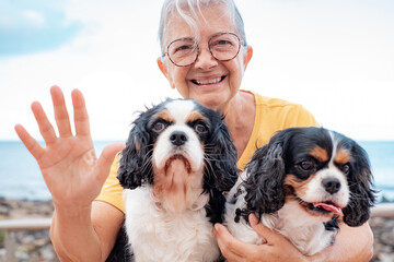 Happy senior woman in yellow jersey sitting close to the beach with her two cavalier king charles dogs looking at camera waving hand . Best friend forever concept