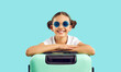 Children's travel. Happy kid teen girl with turquoise travel suitcase on vivid light blue background. Cute funny child girl with two bundles on head and sunglasses is ready for summer vacation abroad.