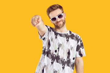 Wall Mural - Happy handsome young man in casual T shirt and sunglasses standing isolated on yellow background and doing pointing finger gesture at camera, at you. Choice, invitation, fashion, party concept