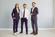 Full length portrait of a group of three young cheerful business people in suits standing confidently, looking at the camera and smiling isolated on a studio grey background. Team work concept.