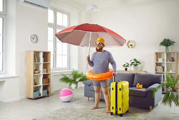 Funny excited chubby man standing at home is finally ready to go on long-awaited summer vacation. Joyful man with suitcase, inflatable circle and large beach umbrella stands in middle of living room.