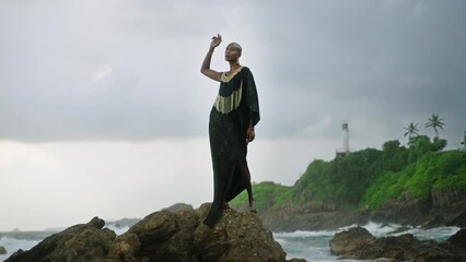 Wall Mural - Gay bipoc man in luxury dress, boutique brass jewelry, makeup poses gracefully tropical rocky hill top on ocean beach. Homosexual ethnic elegant person standing on stone like Oshun orisha. Cloudy sky.