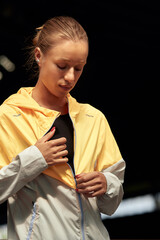 Wall Mural - Young athlete girl getting ready for a workout at the stadium. Attractive slender blonde taking off a yellow-gray sports jacket before training. Active lifestyle, sports and fitness.