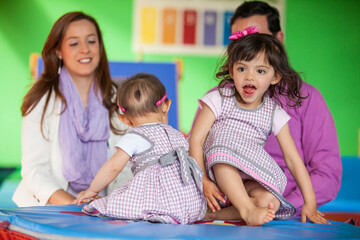 Wall Mural - Beautiful small girl playing with her ten months baby girl sister and their parents on background.. Early stimulation for toddlers and children concept.