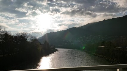 Sticker - Calm river alongside the small village with hills and the sun shining in the background