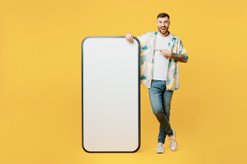 Wall Mural - Full body fun young man he wear blue shirt white t-shirt casual clothes point index finger on big huge blank screen mobile cell phone smartphone with area isolated on plain yellow background studio.