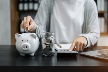 Woman sitting at desk managing expenses, calculating expenses, paying bills using laptop online, making household financial analysis, closer focus on the white piggy bank.