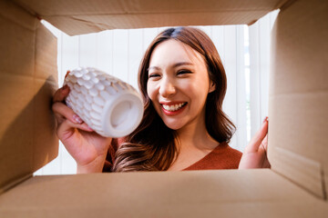 View from inside the box, a Smiling woman unpacking awaited parcel, holding a vase, sitting with a curtain in the background, satisfied happy customer opening a cardboard box with online store order.