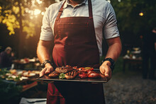 A Man In A Red Apron Holds A Tray With Meat And Vegetables On The Background Of A Barbecue Party.