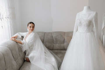 Wall Mural - A wide-angle portrait of a beautiful bride with stylish hair and makeup in a white robe, sitting on a luxurious gray sofa and looking at her wedding dress.