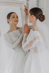 Wall Mural - a beautiful bride in a wedding dress with off the shoulders is standing by the mirror in her room, looking at her reflection in the mirror