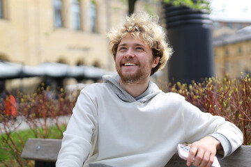 Canvas Print - Smiling curly blond man with a phone in his hands is resting on a park bench.