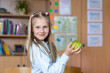 © K-FK - Portrait cute blond confident happy caucasian little blond little kid girl hold hand eating apple in classroom at lunch time pause. Schoolchild in class. Healthy snack food. Back to school concept