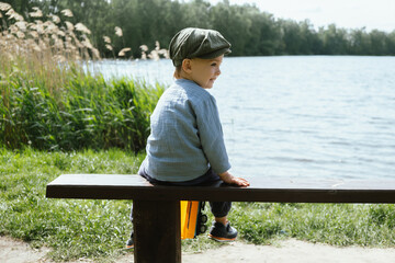 Toddler boy sitting on bench near pond. Little boy enjoys summer walk in countryside. Children outdoor activities. 