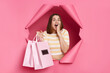 © sementsova321 - Amazed excited woman wearing striped shirt breaking through pink paper hole looking at camera with open mouth holding shopping bags being astonished of visiting mall.