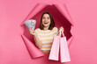 © sementsova321 - Amazed happy cheerful brunette woman wearing striped t shirt posing in torn pink paper wall showing tocamera shopping bags and dollar banknotes cash back.