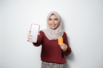 Wall Mural - Cheerful beautiful Asian woman in casual shirt and hijab showing blank mobile phone screen, holding a plastic credit card isolated over white background