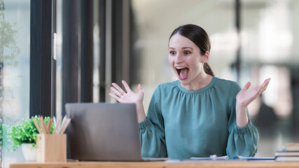 Overjoyed surprised woman looking at laptop screen, sitting at a work desk, reading good unexpected news in email, message, excited by money refund, job promotion, or great sale offer.