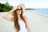 Fototapeta Boho - Portrait of a happy smiling woman in free happiness bliss on ocean beach standing with a hat and sunglasses. A female model in a white summer dress enjoying nature during travel holidays vacation
