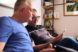 Fototapeta Londyn - Smiling same sex male couple sitting on sofa looking at smart phone together